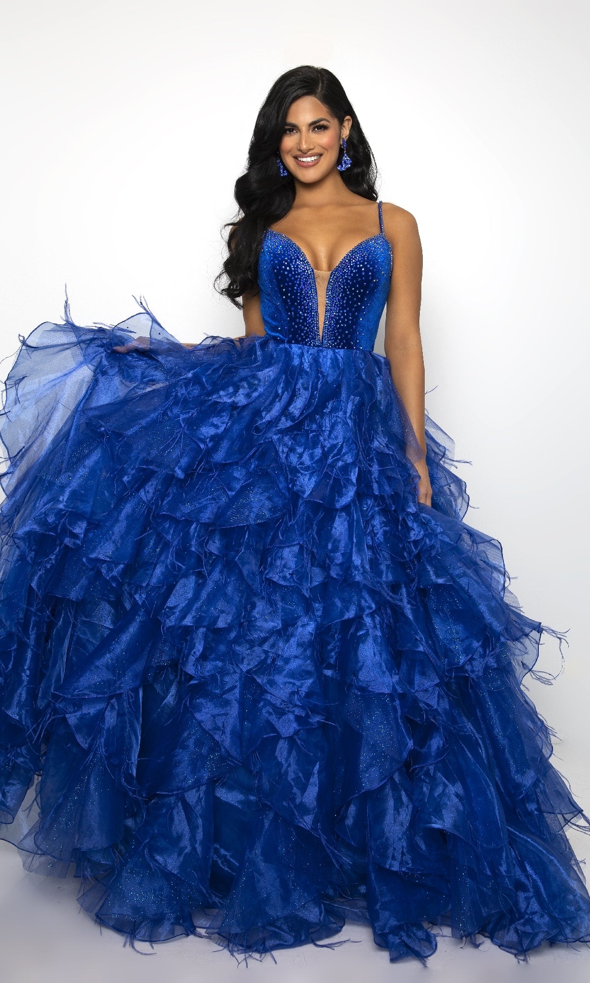A woman wears the Ava Presley 39554 long ruffled prom ball gown in deep blue, featuring a fitted beaded bodice with spaghetti straps, standing against a plain white background.